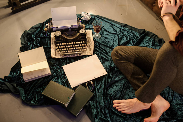 vista lateral del hombre sentado cerca de una máquina de escribir en una tela de terciopelo azul en el suelo en una habitación oscura con bombillas cerca de un sofá de cuero marrón con hojas de papel y libros arround
 - Foto, Imagen