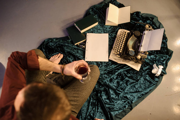 新聞紙や本が並べられた床の上の新聞や濃い青のベルベットの生地にヴィンテージタイプライターの近くに座っている彼の手の中にワインのグラスを持つ男のトップビュー - 写真・画像