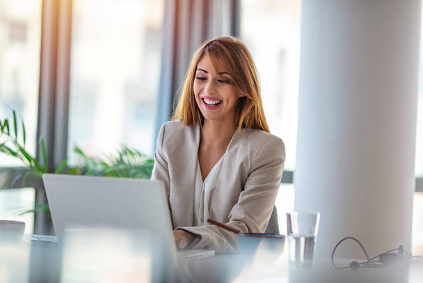 Portrait of a happy businesswoman working at her office desk. Beautiful smiling mixed race businesswoman dressed casual sitting in office and using laptop. - Zdjęcie, obraz