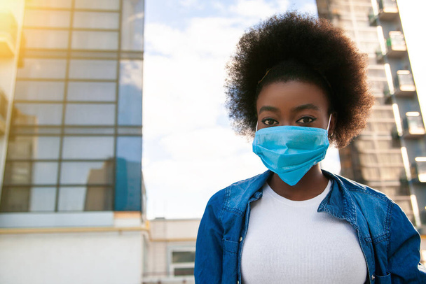 Health, pandemic and safety concept - young american african woman with black power hair, dressed casual, wearing disposable medical protective mask on city background - Photo, image