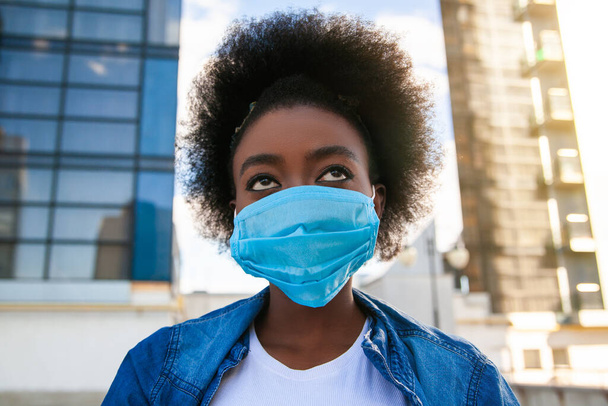 Concepto de salud, pandemia y seguridad: mujer africana americana joven con cabello negro, vestida casual, con máscara protectora médica desechable en el fondo de la ciudad
.  - Foto, Imagen