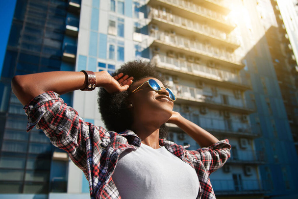 Positive emotions and people concept - young happy black african american female with curly black hair, wearing sunglasses. Over city background. Lifestyle. - Photo, Image