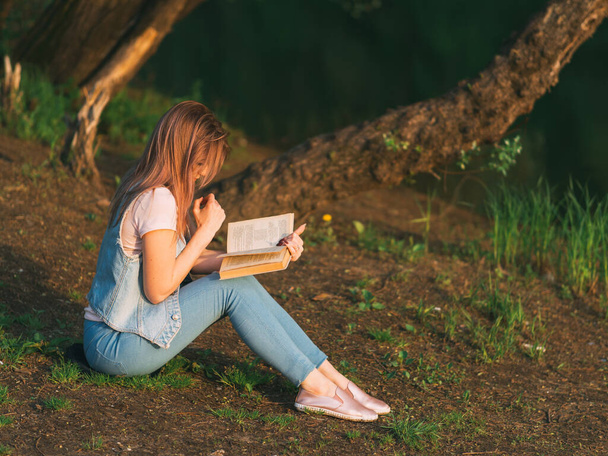 Mladá krásná žena čte knihu při západu slunce u řeky. Přečtěte si knihu v tichosti v přírodě - Fotografie, Obrázek