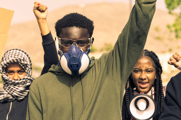 Πορτραίτο ενός νεαρού μαύρου άνδρα με μάσκα αερίων, για πίσω από δύο νεαρές διαφορετικές γυναίκες εθνικότητας, διαμαρτυρία "μαύρων ζωών". Εικόνα - Φωτογραφία, εικόνα