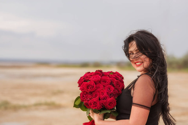 Portrait of a happy girl with long black hair with a large bouquet of red roses on nature. - Foto, Imagen