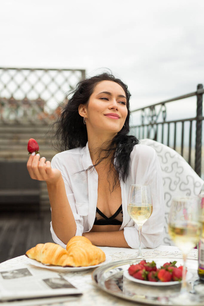 beautiful young woman in a white shirt sits at a festive table with strawberries in hand - Zdjęcie, obraz