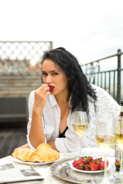 hermosa joven con una camisa blanca se sienta en una mesa festiva con fresas en la mano
 - Foto, imagen