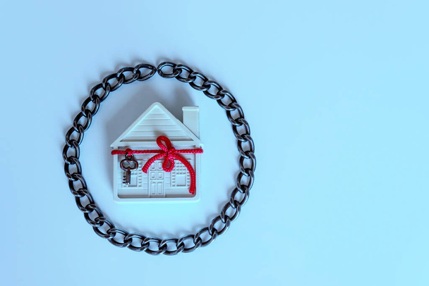  Dom w łańcuchach. Mały biały dom ozdobiony jest dekoracyjnym kluczem na czerwonej wstążce. Koncepcja - ryzyko, utrata mienia, zajęcie, hipoteka.  - Zdjęcie, obraz