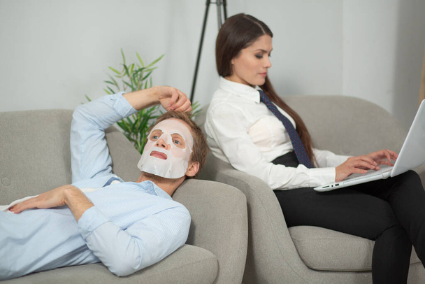 Couple resting on couch using laptop. Young lady working on her computer while sitting on couch at home with her boyfriend laying next to her with sheet mask on - Фото, зображення