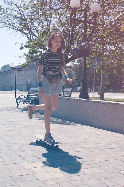 Slender teen girl rides skateboard down an alley in the park in the center of a European city sunny summer day. Instagram or vintage photo - Фото, изображение