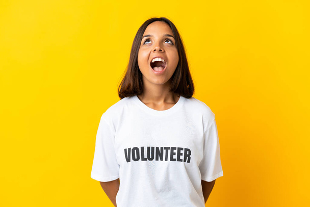 Jonge vrijwilliger vrouw geïsoleerd op gele achtergrond op zoek omhoog en met verrassende expressie - Foto, afbeelding