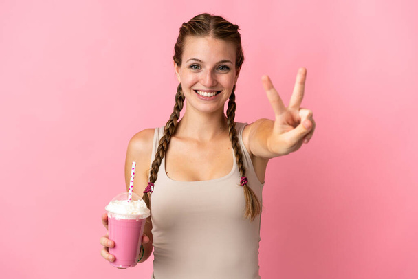 Young woman with strawberry milkshake isolated on pink background smiling and showing victory sign - Photo, Image