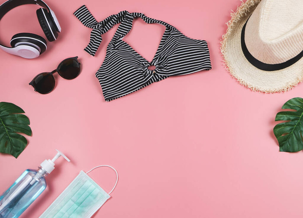 COVID-19 prevention , summer and new normal concept, top view of   bikini swimsuit and women's vacation accessorieswith surgical mask and sanitizer gel  on pink background with monstera leaves. - Foto, Imagem