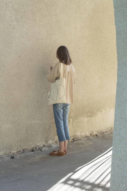 A vertical shot of a female with a white-colored tote bag standing while facing a wall - Zdjęcie, obraz