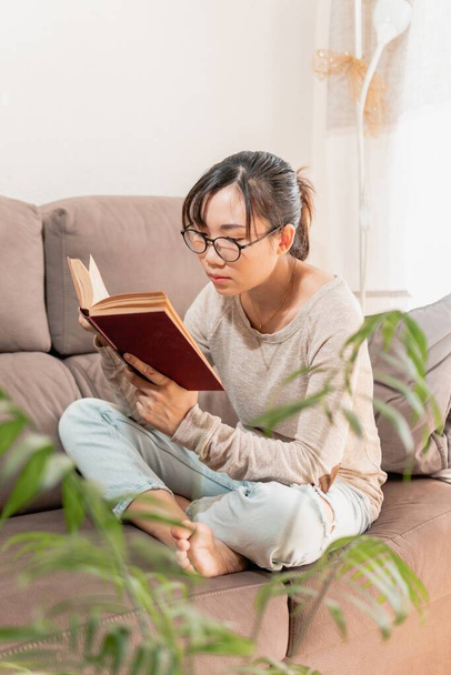 A young Asian woman sitting on the couch and reading a book during Covid-19 quarantine - Foto, Imagem