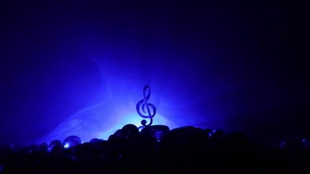 Μουσική έννοια. Μουσικό σύμβολο treble clef από ανοξείδωτο χάλυβα μινιατούρα με πολύχρωμο τονισμένο φως σε ομιχλώδες φόντο. Μουσικά όργανα σε χαμηλό φωτισμό. Επιλεκτική εστίαση - Πλάνα, βίντεο