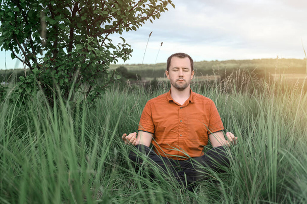 Adult man sitting in the Lotus position and meditating in tall grass. Concept digital detox, connection with nature. - Photo, Image