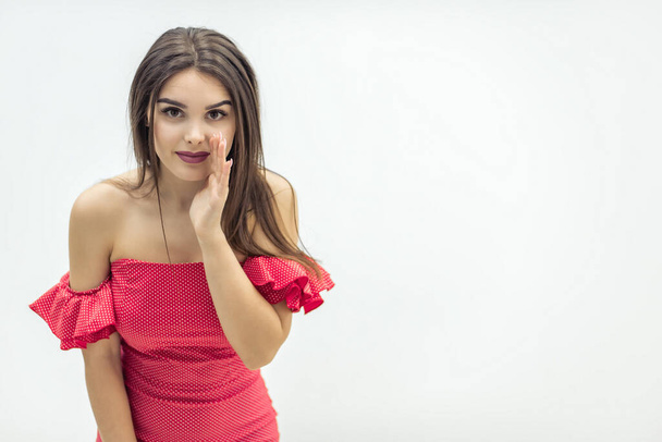 Copyspace photo of beautiful young woman in red dress, wispering some secret to the camera, keeping hand near mouth over white background. - Фото, изображение