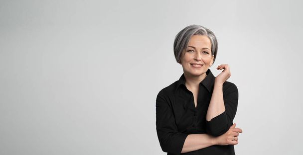 Mature Caucasian woman smiling charmingly with hand near face. Graying elegant woman with short bob hairstyle looks at camera posing on white background. Blank or template with text space at left - Foto, immagini