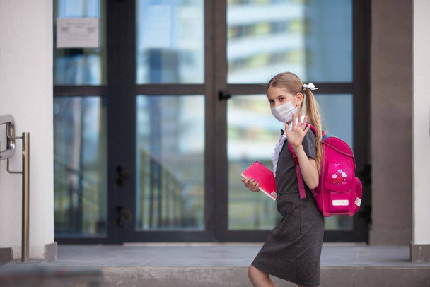 Ein Schulmädchen mit medizinischer Gesichtsmaske und einem rosafarbenen Rucksack steht auf der Treppe ihrer Schule und winkt ihren Eltern oder Freunden zum Abschied zu. Zurück zur Schule. Schutz vor Viren für die Schule.  - Foto, Bild