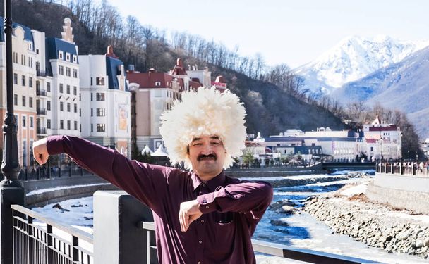 Asian man with a black moustache in dark crimson shirt and in traditional caucasian hat made of white sheepskin is dancing lezginka on a street background. Ski resort Rosa Khutor, Russia. Mzymta River - Photo, Image