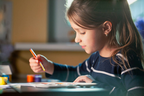 Side view of charming little caucasian smiling schoolgirl writes while sitting at table. Little cute girl draws picture. Concept of teaching children during self-isolation - Foto, Imagen