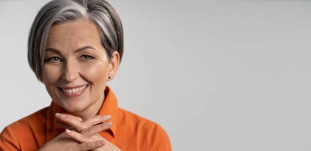 Mature woman broadly smiling with fingers crossed. Happy female model in orange shirt. Close up portrait. Maturity concept. Horizontal template with empty place at right side - Zdjęcie, obraz