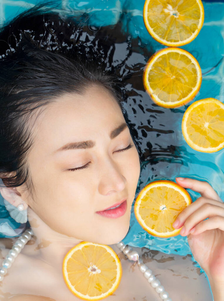 アジアの女性は青いプールで水の上に浮かぶオレンジのスライスで泳いでいて、暑い夏の日を楽しんでいます。美の概念. - 写真・画像