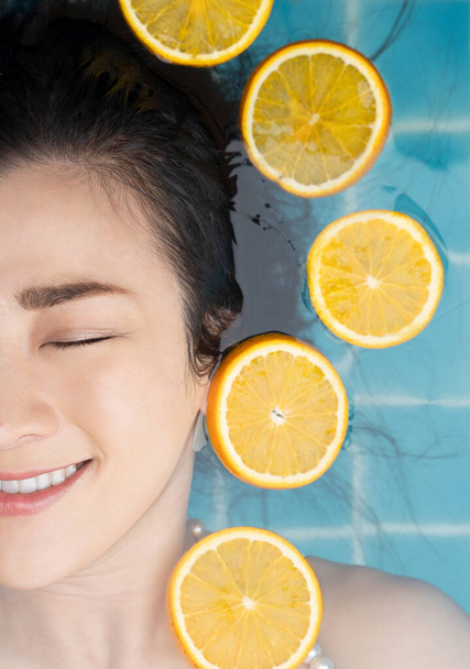 Mulher asiática está nadando com uma fatia de laranja flutuando acima da água em uma piscina azul e desfrutando de um dia quente de verão. conceito de beleza
. - Foto, Imagem