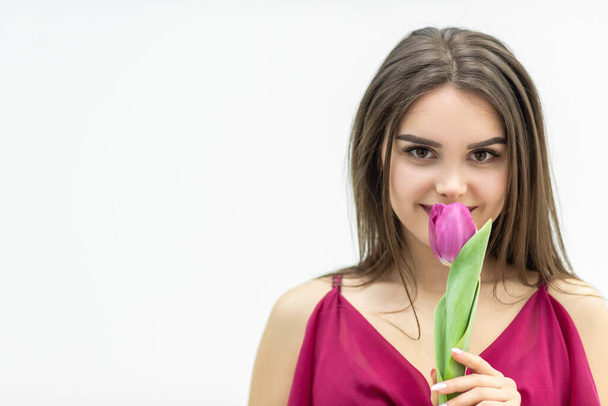 Close up shot of young cute woman smelling pink tulip, slightly smiling at the camera, looking peacefull. - Foto, imagen