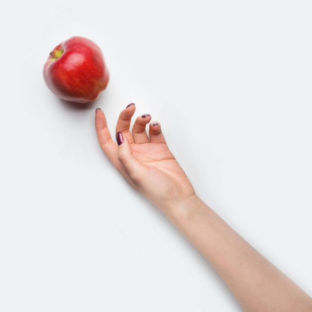 Female hand with red nails dropped red apple on grey background - Фото, изображение