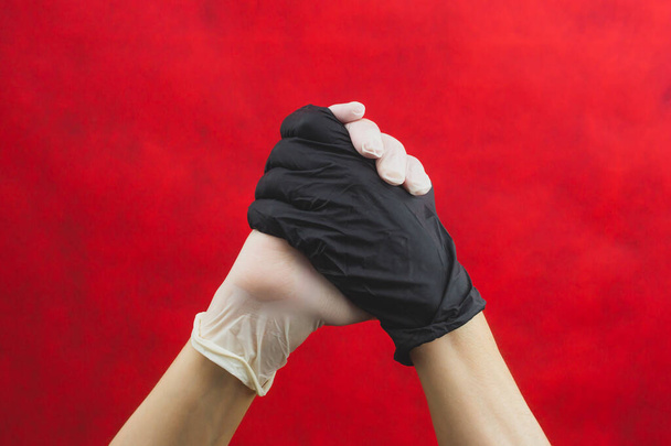 Hands in protective gloves locked together. one hand in black glove, other in white. gesturing hands in medical gloves on a red background - 写真・画像