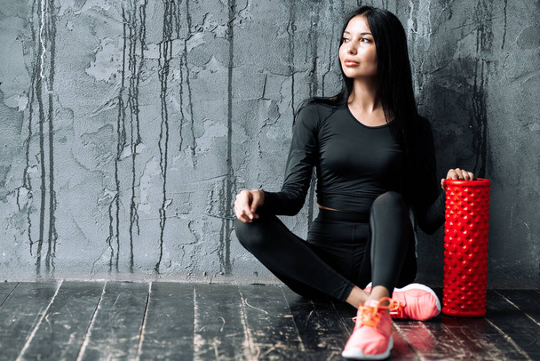 Pilates trainer in a black tracksuit sits on the floor and leans with her back to the wall while holding her hand on a red massage cylinder - 写真・画像