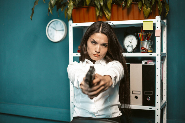 Dangerous woman with gun in office. Crazy or mad angry young businesswoman aiming straight at the camera. Crisis concept. Tinted image - Foto, Imagen
