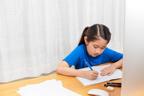 Ασιατικό κοριτσάκι σπουδάζει σε απευθείας σύνδεση μέσω του Διαδικτύου κάθεται και γράφει στο σαλόνι στο σπίτι. Ασία παιδιά γράφουν με μολύβι στο σημειωματάριο. online μάθηση στο σπίτι ή να μάθουν από το σπίτι έννοια. Με χώρο αντιγραφής - Φωτογραφία, εικόνα