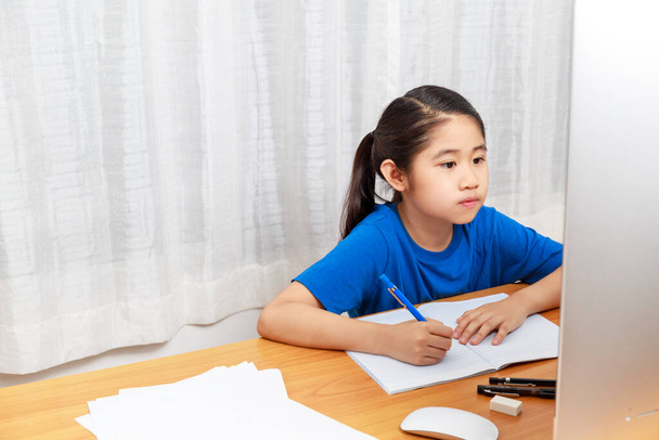 Asiática niña está estudiando en línea a través de Internet sentado y escribiendo en la sala de estar en casa. Asia niños escribiendo con lápiz en el cuaderno. aprendizaje en línea en casa o aprender del concepto de hogar
.  - Foto, imagen