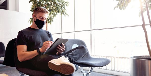 Hombre esperando en el aeropuerto vacío usando mascarilla protectora usando tecnología. - Foto, Imagen
