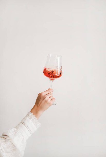 Womans hand in white shirt holding and turning glass of rose wine over white wall background. Wine shop, wine tasting, bar, wine list concept - Zdjęcie, obraz