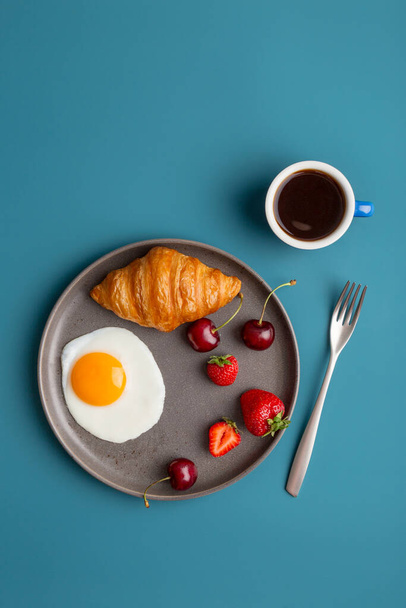 Minimal breakfast at blue background. Fried egg, croissant, berries and cup of coffee at blue backdrop with silverware. Top view, flat lay, copy space. Concept of healthy food for young people - 写真・画像
