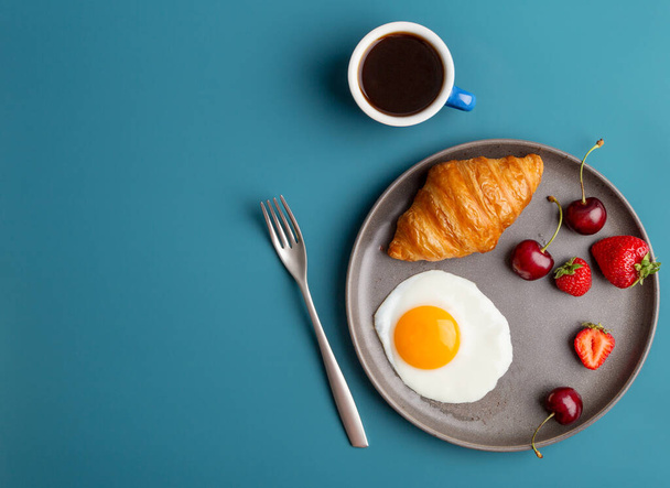 Minimal breakfast at blue background with coffee and croissant with fork and berries. Concept of tasty breakfast. Top view, flat lay, composition of modern food - Photo, image