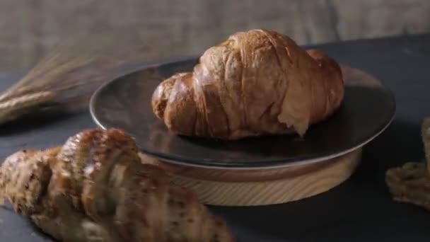 un tradizionale croissant gira su un piatto scuro in cima ad un c-wood rotondo, accompagnato da un altro croissant con cereali, pasta sfoglia e una spiga di grano. Pavimento in ardesia. cibo scuro - Filmati, video
