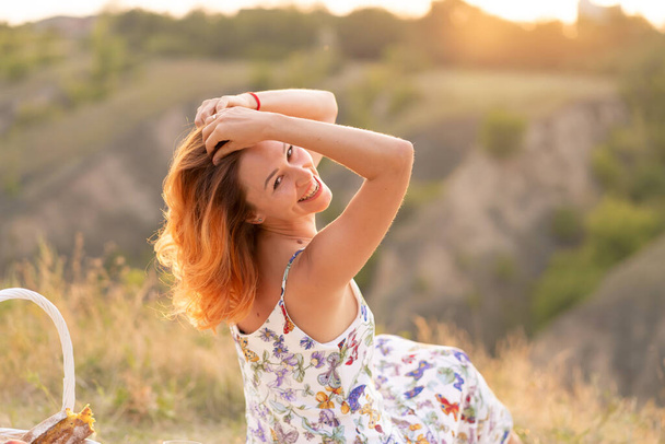 Tender beautiful red-haired girl enjoys the sunset in a field with a hill - Zdjęcie, obraz