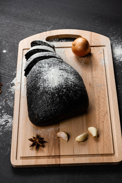 frisches Roggenbrot auf einem Schneidebrett mit mit Mehl bestreuten Gewürzen auf einem schwarzen Holztisch - Foto, Bild