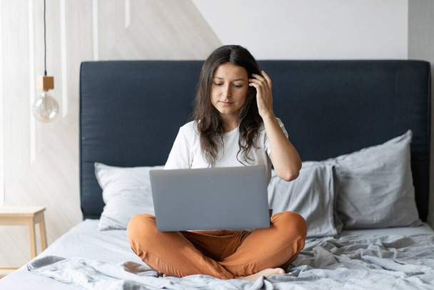 Beautiful young brunette girl with a laptop sitting on the bed. Stylish modern interior. A cozy workplace. Shopping on the Internet. - Foto, imagen