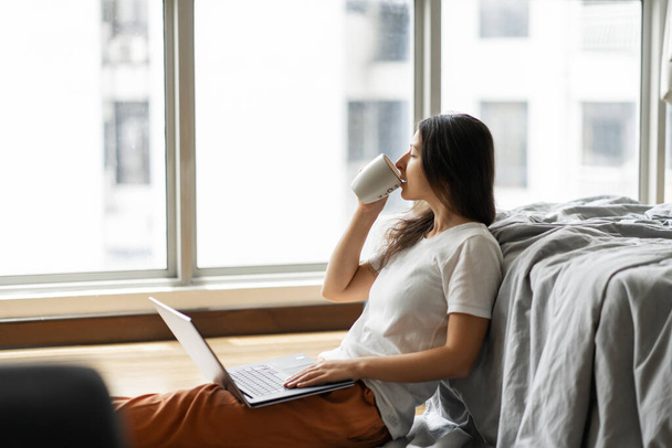 Beautiful young brunette girl working on a laptop and drinking coffee, sitting on the floor near the bed by the panoramic window. Stylish modern interior. A cozy workplace. Shopping on the Internet - Foto, Bild