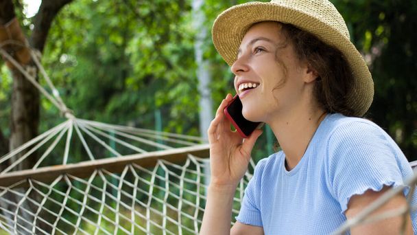 Beautiful young woman talking on mobile phone,relaxing in hammock in green garden park,looking into distance with happy facial expression.Girl in summer hat and blue t-shirt have pleasant conversation - Zdjęcie, obraz
