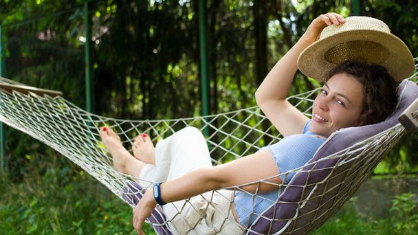 Uśmiechnięta piękna młoda kobieta relaksująca się w hamaku, ciesząca się gorącym letnim dniem w cieniu.Zrelaksowana brunetka trzymająca letni słomkowy kapelusz leżący w humaku w zielonym ogrodzie.Koncepcja weekendowego wypoczynku ummer - Zdjęcie, obraz