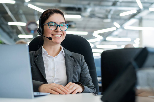 A happy female support operator is sitting at a desk and answering calls. Beautiful smiling woman talking to customers on a headset. Office employee in headphones. - Foto, imagen