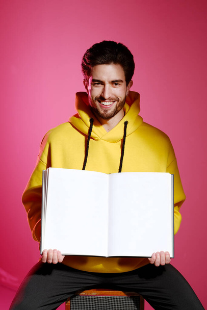 A young man of 25-30 years old in a yellow sweatshirt sitting on an old music column on pink wall background and holding an open book or album.  - Photo, Image