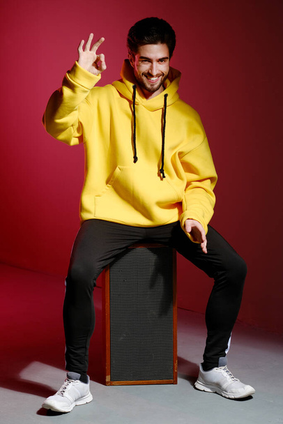 A young man of 25-30 years old in a yellow sweatshirt sitting on an old music column on pink wall background and showing different emotion. - Fotó, kép